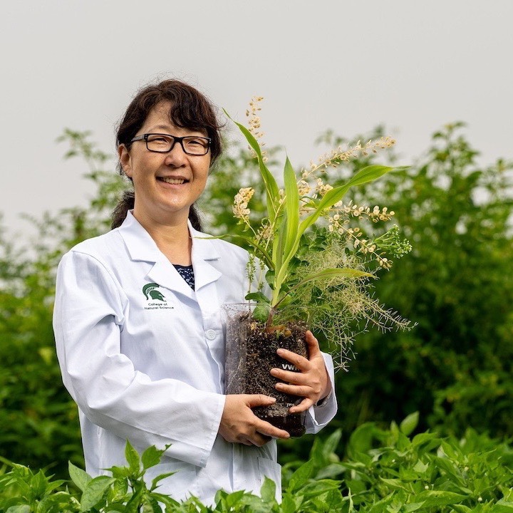 Michigan State University Plant Resilience Institute Director Sue Rhee stands in a field wearing a lab coat and holding a large beaker filled with soil planted with various small crops.