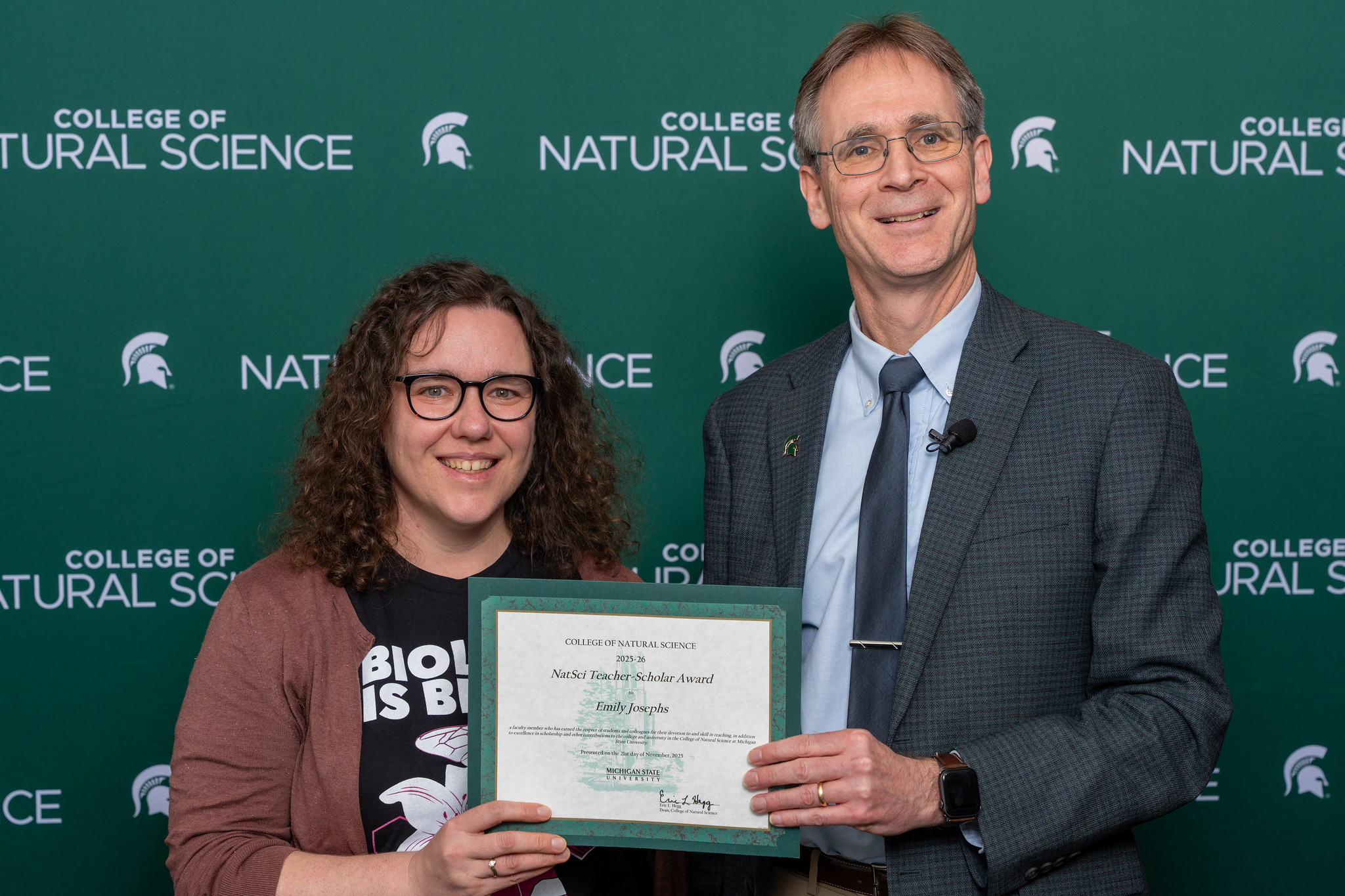 Emily Josephs (left) pictured with Eric Hegg (right) in front of a green backdrop with the MSU College of Natural Science logo while smiling and holding the Teacher-Scholar Award certificate.