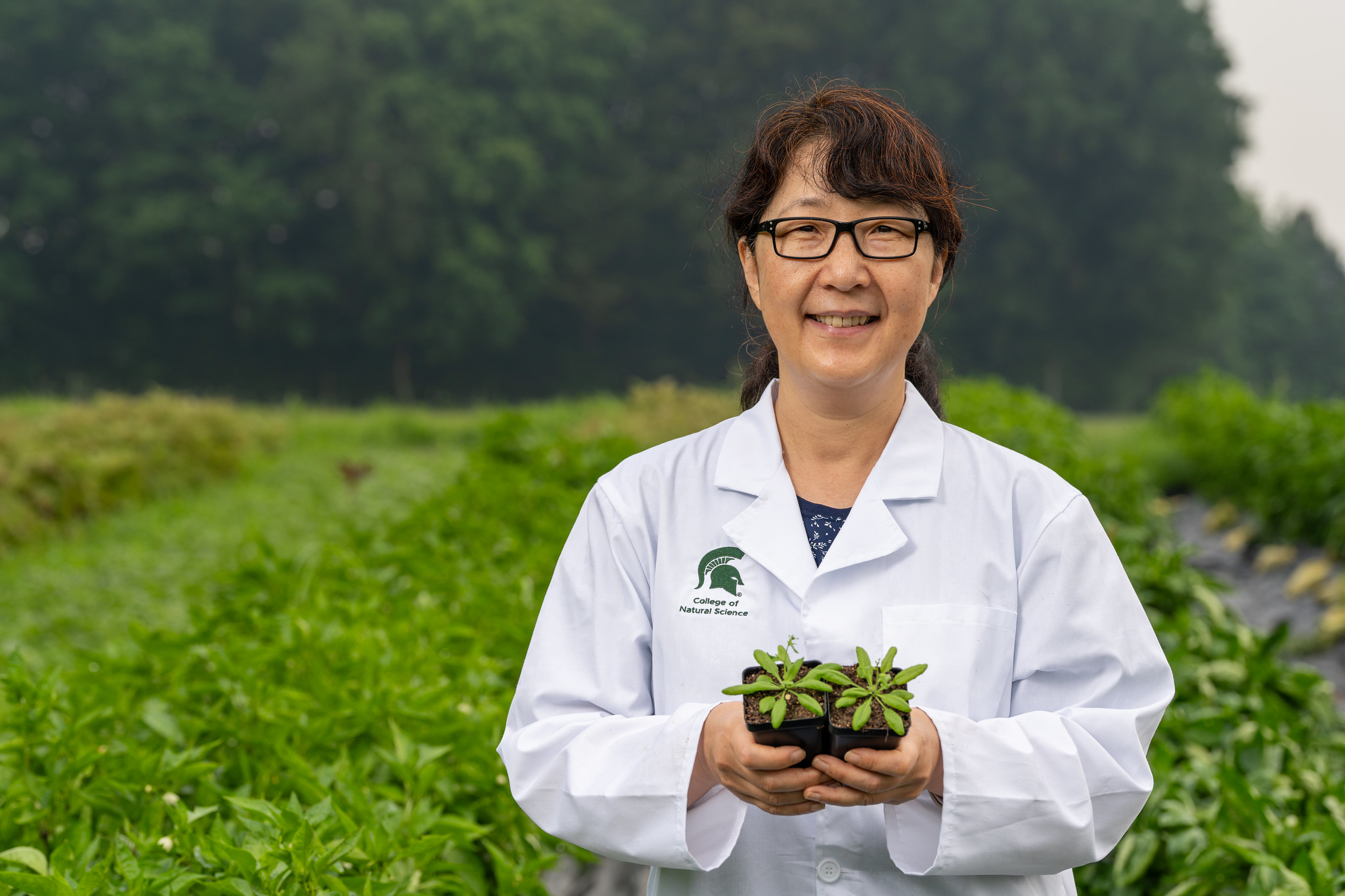 Sue Rhee pictured in a field wearing a lab coat while smiling and holding two small Arabidopsis plants in pots.