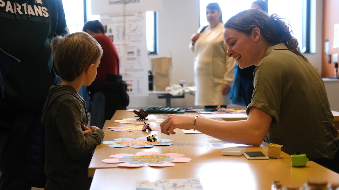 Paige Smith sits at a table across from a child, showing him how bees pollinate flowers with a paper craft on the table.