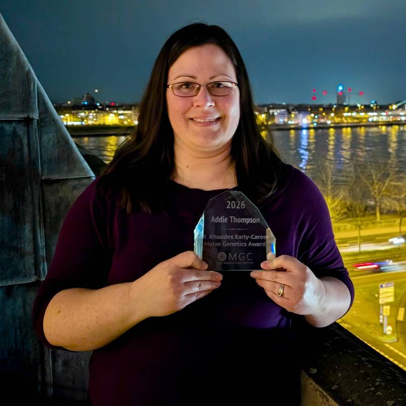 Addie Thompson holds the 2026 M. Rhoades Early-Career Award on a balcony at night overlooking a river.