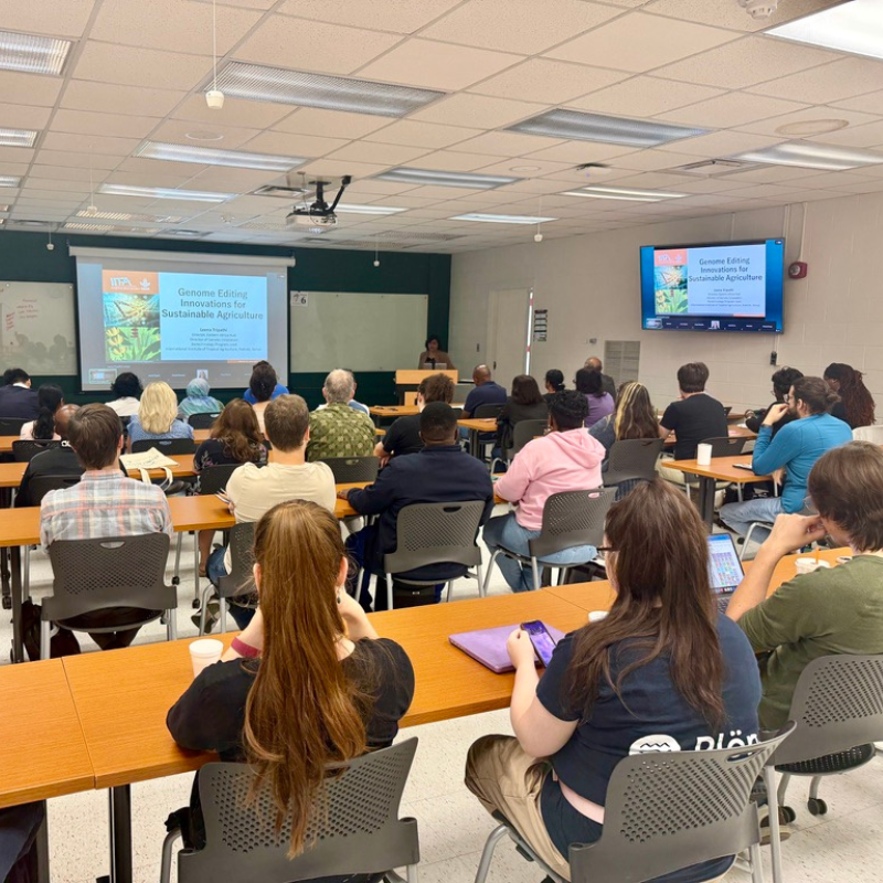 A large group of people sit in rows inside a classroom, and Leena Tripathi stands at a lecturn at the front of the room. Two screens display a slideshow with the following text: 
