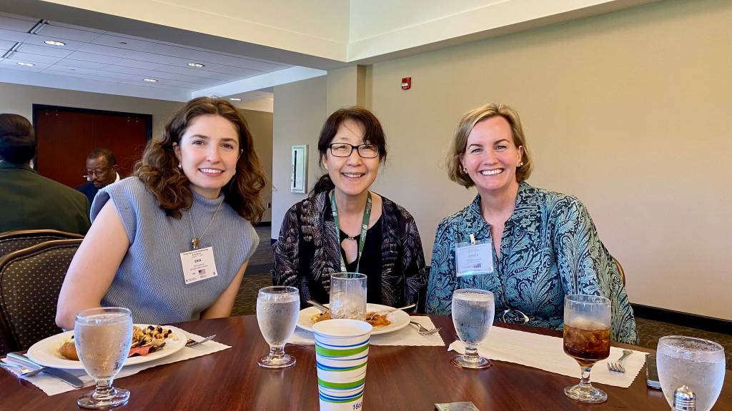 Three women sit at a table together, leaning in and smiling, with plates of food and glasses of water on the table in front of them.