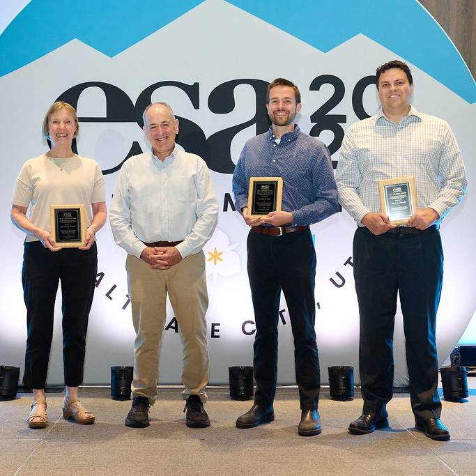 Four people stand together in front of a large sign promoting ESA 2026 while smiling. Three of them are holding small, framed plaques.
