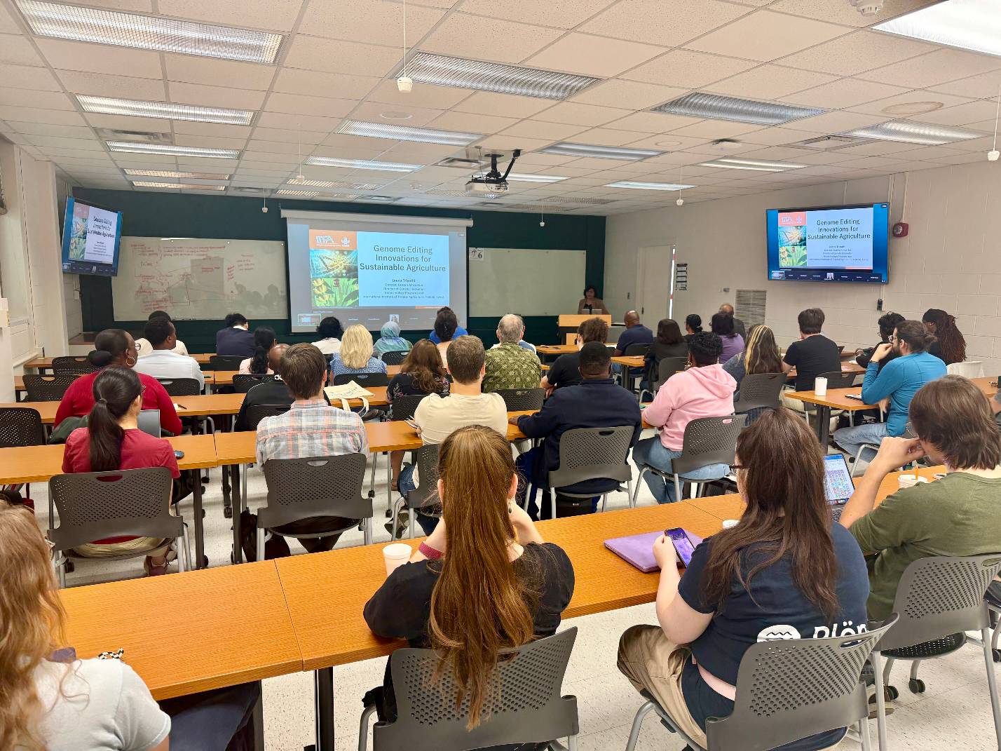 A large group of people sit in rows inside a classroom, and Leena Tripathi stands at a lecturn at the front of the room. Two screens display a slideshow with the following text: "Genome Editing Innovations for Sustainable Agriculture."