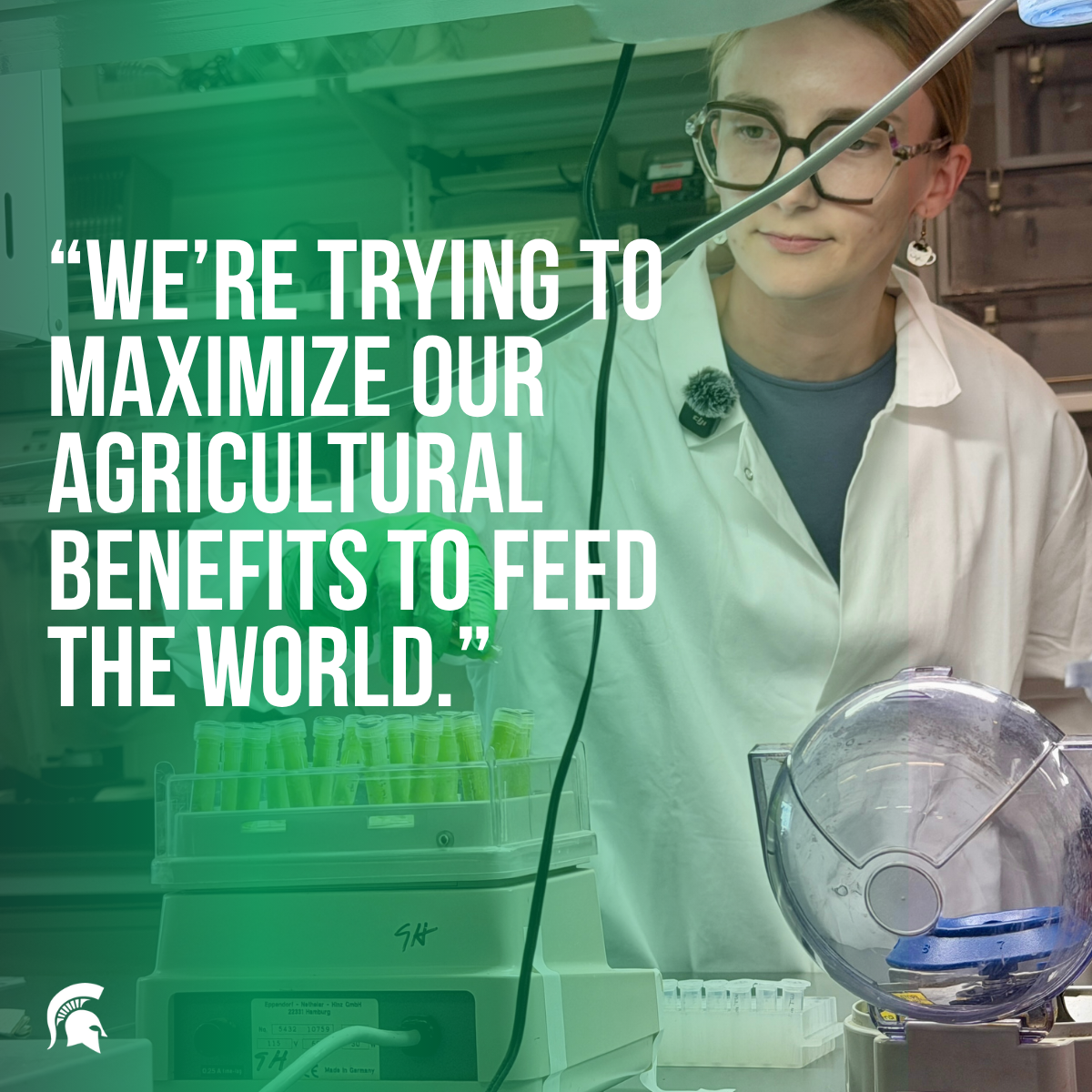 A woman in a lab coat and glasses stands over a lab bench while looking at several small, green test tubes sitting in a shaker. There is a green overlay on the left side with the following text written over it: “We’re trying to maximize our agricultural benefits to feed the world.”