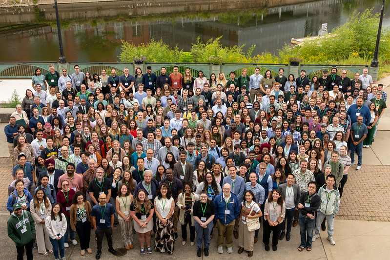 A large group of attendees from the inaugural Great Lakes Plant Science Conference are gathered together outdoors for a photo, with a river and greenery in the background.