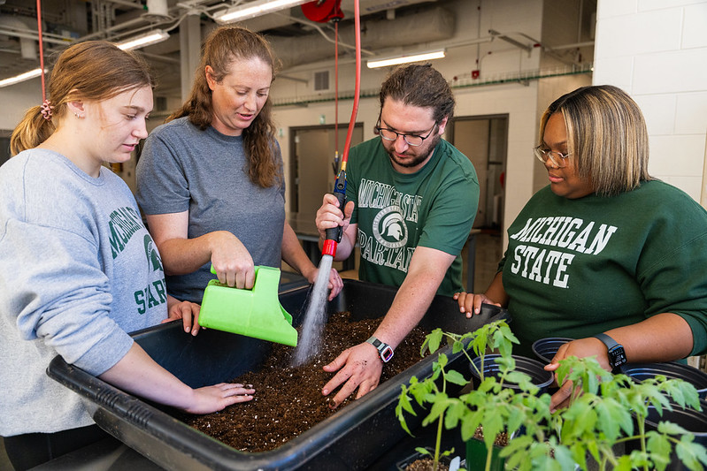 Four people gathered around a large tub of soil next to some potted plants. One of them is holding a hose to water the soil while another is holding a green digging tool.
