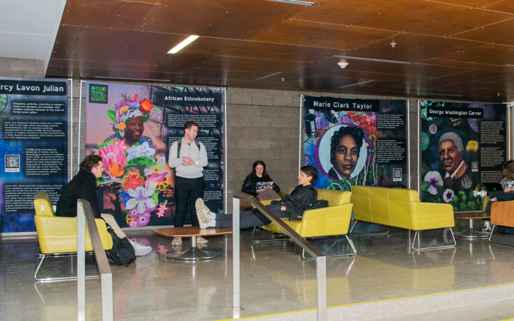 Multiple students sit on couches talking in front of the Plant Cell Atlas exhibit banners featuring art and information on famous Black plant scientists.