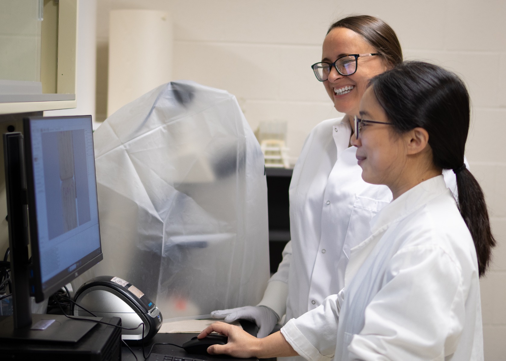 Jinny Yang and Sarah Lebeis working together at a lab computer while smiling and wearing lab coats.