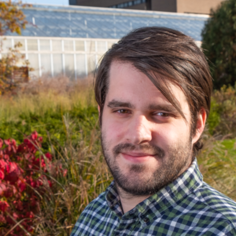 A headshot of Robert VanBuren wearing a flannel button-up shirt outside with various plants and a greenhouse behind him.
