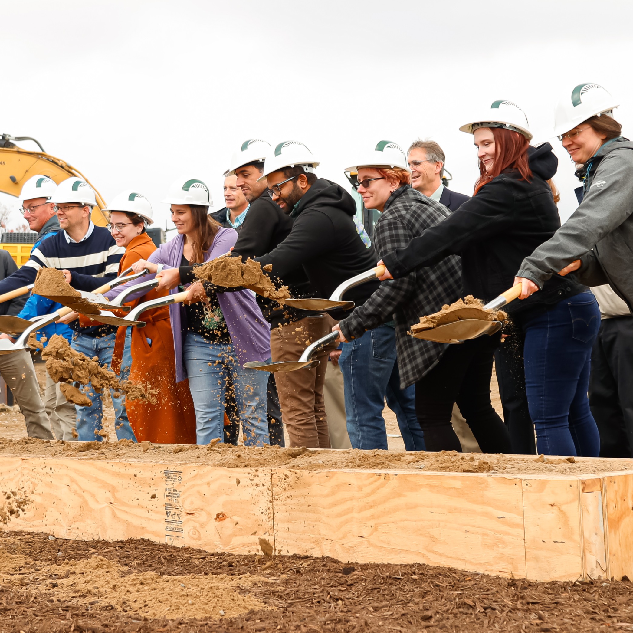 A line of members of the Plant Resilience Institute and MSU-DOE Plant Research Laboratory toss scoops of dirt with shovels. They're wearing Spartan hard hats with construction equipment behind them.