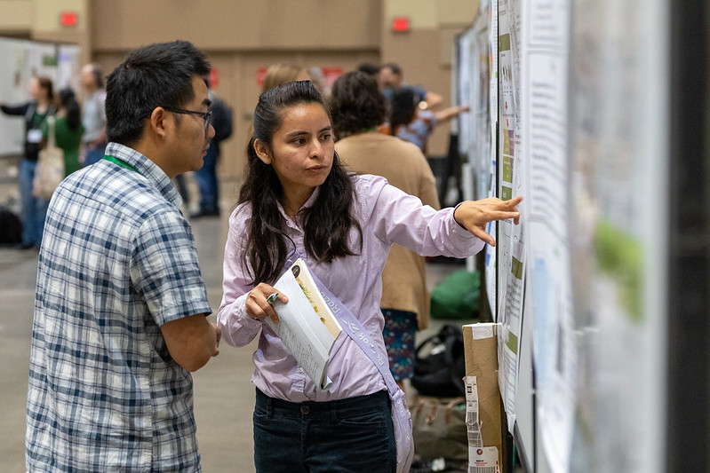 Two individuals are examining a scientific poster. The presenter is pointing at the poster as the other listens attentively. Other poster boards and people can be seen mingling in the background at the First Great Lakes Plant Science Conference.