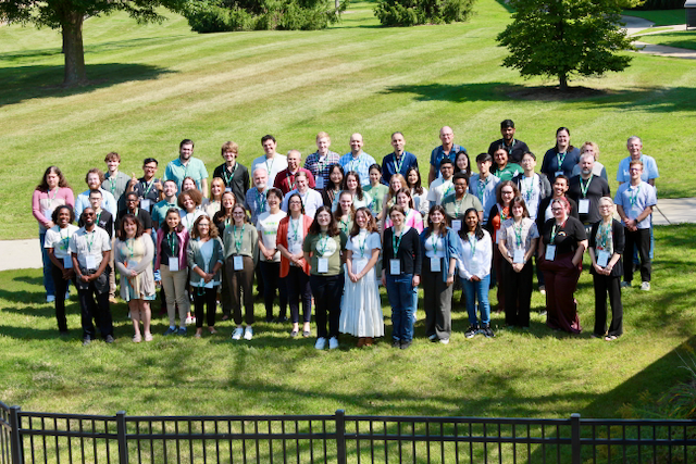 A large group of PRI members wearing lanyards stand for a photo together on a grassy lawn in front of trees at the 2024 PRI retreat.